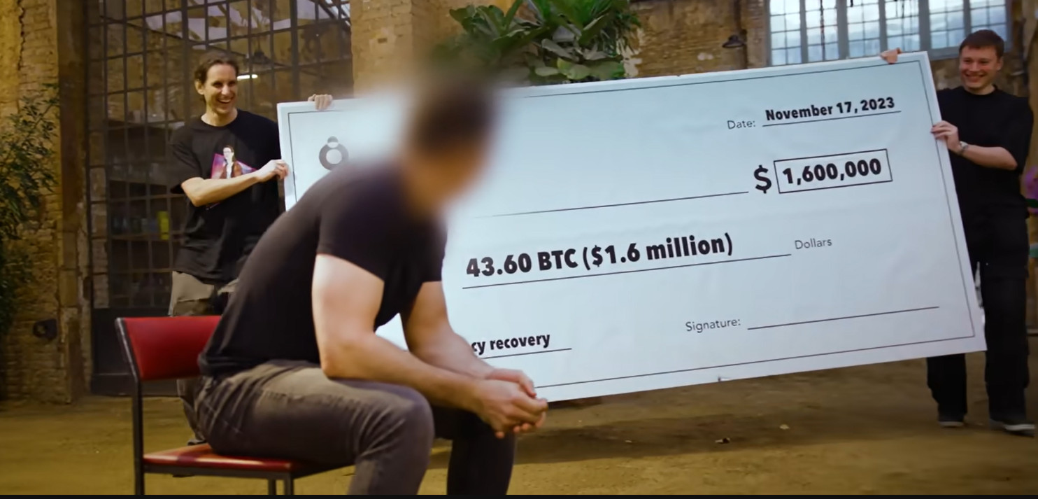 Joe Grand and Bruno presenting a giant check for 43.60 BTC, worth $1.6 million, to a man with a blurred face following a successful hardware wallet recovery.