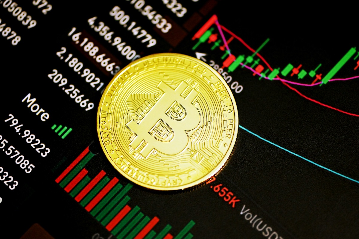 A physical gold Bitcoin coin placed on a digital screen displaying professional financial trading charts and stock market indicators.