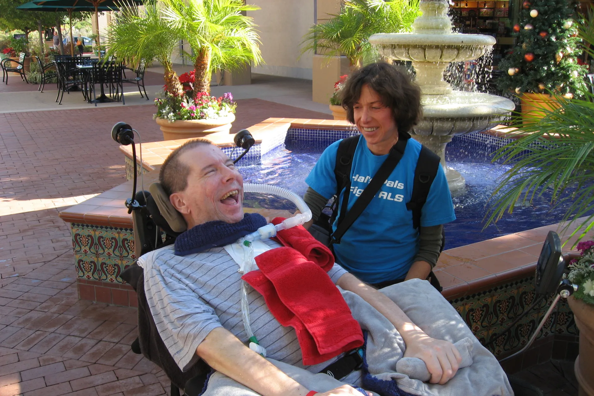A heartwarming photo of Hal Finney in his wheelchair smiling broadly alongside his wife Fran Finney who wears a blue Hal's Pals Fight ALS shirt next to an outdoor fountain, evoking powerful bitcoin nostalgia.